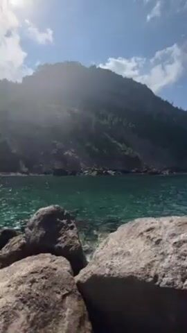 The camera pans slowly to the right to reveal a rocky shoreline at the edge of a clear, turquoise body of water. Large, irregular gray rocks are in the foreground, and behind them the water is calm and clear. A large, steep, and green mountain dominates the left side of the frame, disappearing into a bright, sunny sky with scattered clouds. In the distance, the coastline extends into the horizon. There are no people or other visible objects in the water or on the land. The scene is peaceful and serene, emphasizing the natural beauty of the coastal environment.