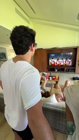 A group of young adults are watching a football game on television in a living room. Two are lying on a large sofa while others stand around the room. One man walks towards the sliding glass doors leading to an outdoor area. The room has wood paneling and white walls and ceilings. There are food and drinks on the table in front of the sofa.