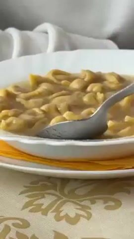 A close-up shot shows a white bowl filled with tortellini in broth, resting on an orange napkin. A spoon is shown scooping some of the tortellini. The bowl sits on a plate with a floral pattern. In the background, there's a white fabric draped.