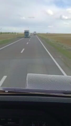 A dash cam video shows a highway with several trucks and cars. The road is paved and has white lane markings. The surrounding landscape is flat and covered with dry grass. The sky is cloudy. In the foreground, a roll of paper towels is visible on the dashboard. The video appears to be taken from inside a vehicle.