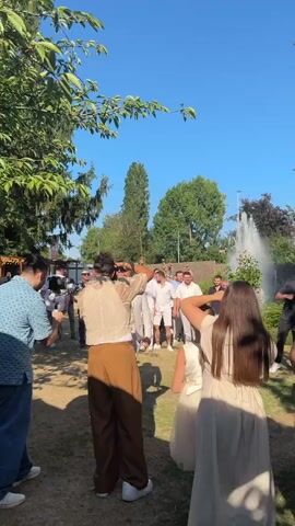 The video shows a group of men in a backyard on a sunny day, possibly at a wedding. In the foreground, a man in a beige suit with a bowtie and a red boutonniere is holding a selfie stick. Behind him, a group of men in white shirts and shorts are gathered, some appearing to be posing for photos. A woman in a light-colored dress is also visible in the foreground, facing away from the camera. Trees and foliage are present, and a fountain is visible in the background. The overall atmosphere appears to be festive.