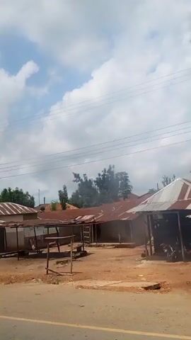 The video captures a moving shot from a vehicle, looking out at a street scene. The camera pans right, revealing various buildings and infrastructure. In the foreground, a motorcycle with two occupants is visible. The surroundings consist of dirt roads, buildings with corrugated metal roofs, and concrete structures. A solar-powered street light stands prominently on the right side of the road. The sky is partly cloudy. As the camera continues to pan, more buildings come into view, including what appears to be a gateway with decorative pillars. Further along, multi-story buildings with green roofs are seen. The general environment suggests a developing urban or semi-urban area.