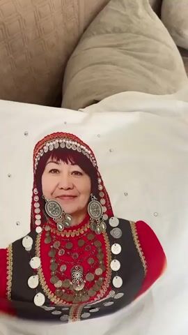 A video shows a close-up of a white fabric, likely a pillowcase or sheet, featuring a printed image of a woman. The woman is depicted wearing traditional clothing, including a red head covering adorned with small silver circles, elaborate layered necklaces made of coins and metal pieces, and large circular earrings. She is also wearing a dark vest over a red garment. The print is detailed and colorful, with small sparkling gems scattered around the image on the fabric. The background behind the fabric shows blurred textures of what appears to be soft furnishings, possibly a bed with pillows. The camera slowly pans or slightly zooms, offering different angles of the printed image and its surroundings.