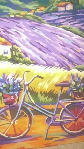The video shows a close-up, side-view of a painting. The painting depicts a rural landscape scene with a prominent lavender field stretching into the distance. In the foreground, a bicycle is parked on a dirt path. The bicycle is a light purple color with baskets on the front and back filled with purple flowers. The wheels of the bicycle have orange spokes. The background features rolling green hills, scattered houses with red roofs, and cypress trees. The sky is painted with soft, blended colors of blue, purple, and pink. The painting style is characterized by bold, flat areas of color and simplified shapes. The texture of the wall behind the painting is visible.