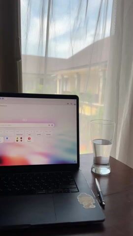 A laptop rests on a wooden desk next to a half-full glass of water and a pen. A sticker is affixed to the laptop. The laptop screen is open, displaying a web browser with various icons and text in a non-English language. A sheer white curtain hangs behind the desk and laptop, partially obscuring the view outside the window. Through the curtain, a multi-story building with a brown roof and a swimming pool with gazebos can be seen. The sky is visible above the building. The camera zooms into the curtain and the window to view the scene outside more clearly.