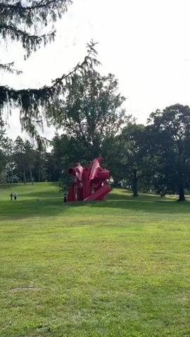 A wide shot captures a vibrant red abstract sculpture situated on a lush green lawn. The sculpture, characterized by its tubular and curved forms, dominates the center of the frame. In the background, a line of trees forms a natural backdrop, with sunlight filtering through their leaves. To the left of the sculpture, two individuals are visible, appearing to be walking or standing on the grass. The overall environment suggests an outdoor setting, possibly a park or sculpture garden, under a bright sky.