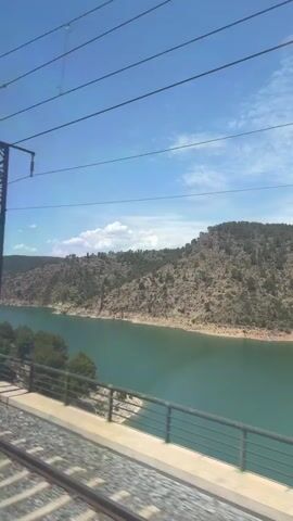 This video shows a view from a moving train crossing a bridge over a body of water. The water is a deep teal color and appears to be a lake or reservoir. On the far side of the water, there is a rocky, tree-covered hillside. The sky is blue with scattered white clouds. Overhead, there are power lines and a support pole for the overhead electrical system for the train. The train tracks are visible in the foreground, and the side of the bridge with a metal railing is also visible. The scene is brightly lit by sunlight, indicating it is daytime. The video is a continuous shot, showing the scenery passing by as the train moves. There are no people visible in the video.
