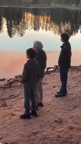 Three people stand on a sandy shore overlooking a calm lake, their backs to the camera. The person on the left appears to be a young boy in a grey jacket and jeans. In the middle stands an adult with grey hair, wearing a dark jacket and tan pants. On the right, a taller individual is dressed in a dark jacket and jeans, with headphones around their neck. The lake reflects the trees on the opposite shore and the sky, which has a soft pink hue. The shoreline consists of sand with a few pieces of driftwood. A building can be seen in the distance on the far shore. The lighting suggests either dawn or dusk.