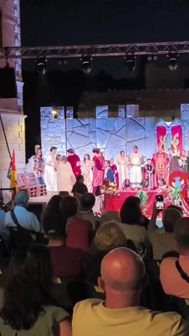 This is a vertical video of an outdoor evening theatrical performance. The stage is set with a backdrop resembling ancient stone walls, adorned with red and gold banners. A crowd of people is seated in the foreground, facing the stage, some holding up their phones to record or take pictures. On stage, a large cast of performers in period costumes, mostly in rich reds, whites, and golds, are gathered. A central group of performers appears to be engaged in some sort of ceremonial or dramatic presentation. There are tables on stage with fruit and other decorations. Stage lighting is visible overhead, illuminating the scene. A flag with yellow and red colors is visible on the left side of the stage. The overall atmosphere suggests a historical reenactment or a play.