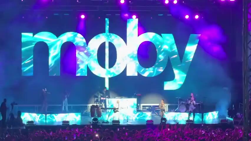 A live concert performance is shown, with the word "moby" displayed in large white letters against a vibrant purple and pink backdrop. The letters are filled with a colorful, abstract, pixelated pattern. Below the logo, a band performs on a raised stage, illuminated by bright stage lights and smoke effects. Several musicians are visible playing instruments, including a guitarist, a drummer, and keyboardists. The audience in the foreground is a blur of light and color, suggesting a large crowd. The overall atmosphere is energetic and festive.