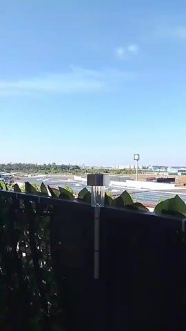 The video shows a view of a bright, sunny sky with a few wispy clouds. In the foreground, there is a dark fence with artificial green leaves attached. A solar-powered garden light with a silver top is mounted on the fence. Beyond the fence, a flat, expansive area with what appears to be solar panels is visible. Further in the distance, a line of trees and buildings can be seen, with some taller structures forming a city skyline. A small dark object, possibly an airplane, is visible in the sky. The camera appears to be handheld and pans slightly to the right.