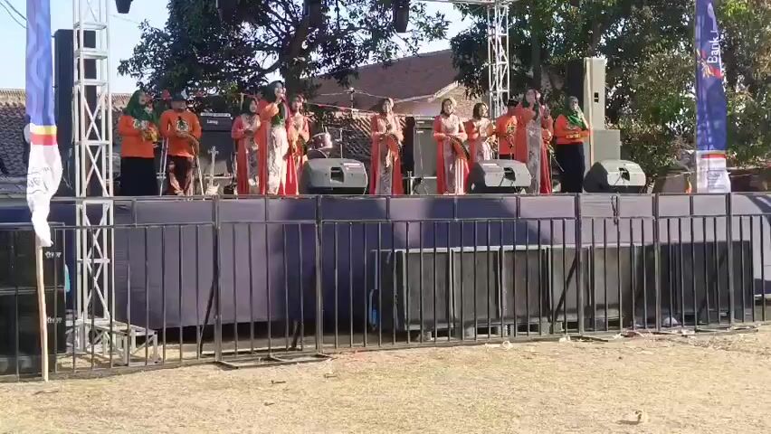 A group of women on a stage. The women are wearing orange tops and patterned dresses. Most of them are also wearing headscarves. Some of the women are holding tambourines. They appear to be performing for an audience, although the audience is not visible in the frame. The stage is set up outdoors, with trees and buildings visible in the background. There are speakers and lighting equipment on the stage. The ground in front of the stage is covered in dry grass. There are banners on either side of the stage, one of which has text that is too small to read. The overall impression is of an outdoor performance or festival.