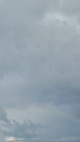 A short clip of a cloudy sky, with a rainbow appearing in the lower right corner. As the camera pans down, the rainbow becomes more prominent and a rooftop with some greenery and electrical wires comes into view.