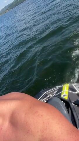 A man is riding a jet ski on a body of water. The water is dark blue and choppy, with white foam trailing behind the jet ski. The sky is a bright blue with a few white clouds. In the background, there are green hills covered with trees. The man is facing the camera and looking serious. He has short, dark hair and is shirtless. The jet ski is black with yellow and white accents. His legs are visible in the foreground, one of which has red marks on it.