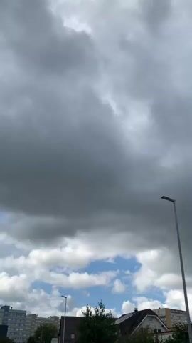 The video shows a time-lapse of a cloudy sky over a suburban landscape. Several buildings and trees are visible below the sky. A street light is prominently featured in the foreground. The clouds are dense and dark, suggesting an impending storm. The sky transitions from mostly overcast to partly cloudy with patches of blue sky appearing as the video progresses.