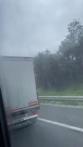 The video shows the rear of a white semi-trailer truck with red trim on a road. The truck has a license plate that reads "CB 6752 EA". There are speed limit stickers on the back of the trailer, indicating 50, 80, and 90. The truck is branded with "SCHMITZ". The view appears to be from another vehicle traveling alongside or behind the truck, as evidenced by the slight tilt of the camera and the reflection of a window frame in the foreground. The weather appears overcast, and trees line the right side of the road, separated by a metal guardrail. The road surface appears wet.