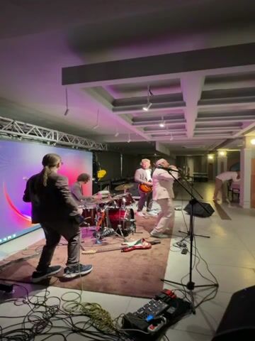 A live band performs on a stage in front of a large screen displaying a colorful abstract image. The band consists of a drummer, two guitarists, and a singer. They are all dressed in suits. The singer wears a light pink suit and a head covering, while the other band members wear darker suits. The stage is set up on a rug, with musical equipment like guitars, amplifiers, and microphones visible.

The scene appears to be an indoor event, possibly a party or reception, with tables and chairs set up in the background, suggesting a seated audience. Several people are seen dancing on the dance floor in front of the stage, with one woman in a black dress spinning joyfully. The room is lit with a mix of ambient lighting and spotlights directed at the stage, creating a vibrant atmosphere. The ceiling has a distinctive coffered design. The overall mood seems festive and energetic.

There is no visible text in the media file. The location context provided, Каджи-Сай 2, Kyrgyzstan, suggests the event is taking place in that region. The time of day is likely evening or night, given the indoor lighting and the nature of a live band performance at an event.