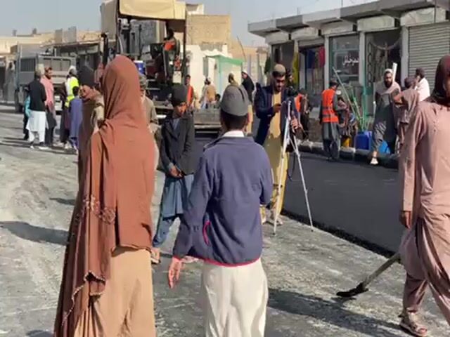 The video shows a street in what appears to be a Middle Eastern or South Asian town. A number of people are walking along the street, including men, women and children. Many of the men are wearing traditional clothing, including hats. One woman is wearing a brown burqa. Road work appears to be in progress. A vehicle is involved. A man is filming with a camera on a tripod. The weather looks sunny.