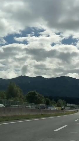 The video shows a view from a car driving on a highway. The highway has multiple lanes and concrete barriers. There are other vehicles on the road, including cars and trucks. On the left side of the highway, there is a fence and some trees. In the background, there are mountains covered in trees. The sky is cloudy with some patches of blue. The video appears to be shot during the daytime.