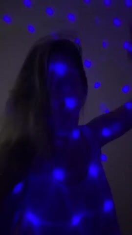 A medium shot shows a woman with blonde hair, wearing a white sleeveless top, dancing in a dark room. Blue disco lights project onto her and the wall behind her. She raises her arms to head height and moves her hands around. She holds her right hand in a fist with her index finger and pinky extended. She then lowers her hands.