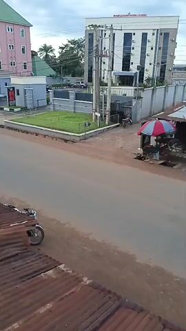 This is a video of a street scene in Achalla, Nigeria. The video captures an urban setting with several buildings of varying architectural styles. On the left side of the street, there is a tall, pink building with multiple stories and windows. Adjacent to it is a smaller, modern grey building with a glass facade, possibly a reception area or office. Across the street, a larger, white building with a modern design stands prominently. It features numerous windows and appears to be a hotel or commercial establishment, with "ROYAL LUXURY HOTEL & SUITES" visible on its facade.

The street itself is paved with asphalt and shows signs of wear. In the foreground, a rusty corrugated metal roof is visible, suggesting a lower-income structure or older development contrasting with the newer buildings. A motorcycle is parked on the side of the road. On the right side of the street, a small stall with a red and blue umbrella is set up, likely a vendor's stand, with goods and a person visible beneath it.

The scene appears to be during the daytime, with overcast skies suggesting cloudy weather. The overall impression is of a developing urban area where modern infrastructure coexists with older, perhaps more traditional, structures. There is no obvious activity or event taking place other than the general presence of buildings, vehicles, and a street vendor. The presence of a hotel suggests it's a place where visitors might stay, implying a degree of commercial activity.