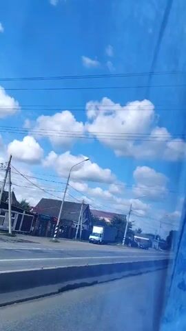 The video shows a view from a moving vehicle looking out at a roadside scene. The sky is bright blue with scattered white clouds. A road is visible with cars and trucks driving on it. On the side of the road, there are buildings, trees, and streetlights. There are also power lines running across the scene. The perspective is from a window that appears to be tinted or dirty, as there is a bluish hue to the image. The video is short and appears to capture a fleeting moment of the journey.