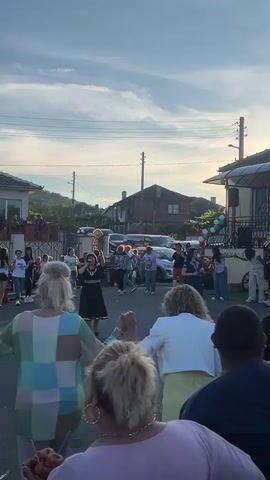 A group of people are holding hands and dancing in a circle in the street. The sun is setting in the background, casting a warm glow on the scene. Some of the people are women, and they are all dressed in casual clothing. There are houses and cars in the background, suggesting that this is a residential area. The atmosphere appears to be festive and celebratory.