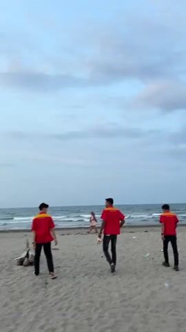 The video shows a beach scene with the ocean in the background. Several people are visible on the sandy beach. In the foreground, two young men in red shirts and black pants are walking away from the camera. Another person in a red shirt is visible further back, and behind them, a person in a light-colored outfit is walking towards the water. In the middle ground, there is a motorcycle and a small wooden structure. The sky is cloudy, and the lighting suggests it is either early morning or late afternoon. The waves are visible in the ocean.