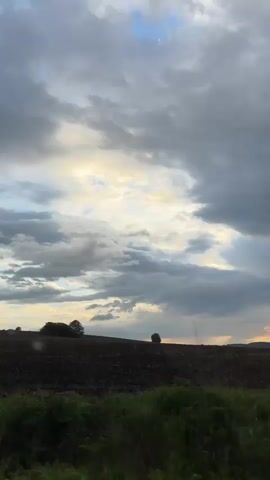 The video shows a view from a moving vehicle, presumably a car, traveling through a rural landscape. The sky is predominantly overcast with a mix of dark grey and white clouds, though there's a patch of blue visible higher up. Hints of sunset colors peek through the clouds along the horizon, creating a golden glow. The foreground features fields, some appearing freshly plowed with dark soil, and patches of green vegetation. Trees are interspersed throughout the scenery, their forms silhouetted against the sky. The movement suggests the video was taken while the vehicle was in transit, giving a blurred impression of the landscape passing by.