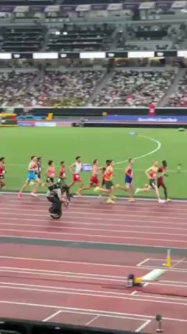 A long shot shows several runners competing in a steeplechase event at a stadium. All are male, dressed in uniforms representing different countries, and are running in the same direction across a red track. A cameraman on a Segway follows the runners. The racers jump over blue and yellow barriers. In the foreground there is a man kneeling with his back to the camera, and equipment beside him. The stadium is full of spectators. International flags line the upper edges of the stadium.