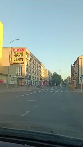 This media file captures a view from a car driving down a street in Gryfino, Poland, during what appears to be late afternoon or early evening due to the warm, low-angled sunlight casting long shadows. The sky is clear and a light blue, suggesting pleasant weather.

The scene depicts a typical urban street lined with multi-story apartment buildings and commercial establishments on the ground floor. The buildings are predominantly light-colored, with numerous windows and balconies visible on the residential floors. The ground-level shops have signs, but many are too small or indistinct to be read clearly.

On the left side of the street, a prominent yellow billboard is visible on a building. It displays text in Polish, with a large "1.1 km" indicator and the slogan "WŁĄCZAMY NISKIE CENY!" (We are turning on low prices!). Below this, "mediaexpert" is clearly legible, suggesting an advertisement for an electronics store. Further along the street, other storefronts are visible, including what appears to be a pharmacy with green signage.

Several people can be seen walking along the sidewalk and near the shops, and a few cars are present on the road, indicating moderate activity. The road itself is paved with asphalt and features pedestrian crosswalks and lane markings, typical of a city street. In the distance, trees and more buildings continue to line the route. The overall impression is one of a calm, everyday urban environment in a Polish town.