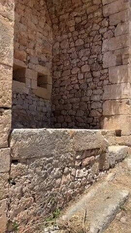 The video shows a stone fortress. The camera starts close to a corner of a stone structure, then pans up and forward along a stone path that runs between arches to a stone courtyard with several doorways. The sky is blue with few clouds. The ground is dry and covered in dry grass and weeds. The stonework is aged and weathered.