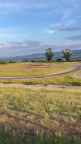 The video presents an expansive view of an outdoor go-kart track located in Anevo, Bulgaria, under a partly cloudy, late afternoon sky. The track, constructed from asphalt or concrete, features several turns and winds through predominantly green, slightly overgrown grassy areas. Two prominent trees stand near the track, and a small pond is visible next to one of the turns. In the background, a range of mountains stretches across the horizon. A go-kart is seen driving along the track, traversing from the left side of the frame towards the right. The late afternoon sunlight casts a warm, soft glow on the landscape, contributing to a serene atmosphere. There is no visible text in the video.