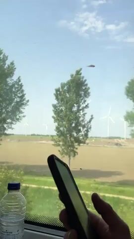 A hand holds a black smartphone in front of a train window. Outside the window, trees, fields, and distant windmills are visible as the train moves. A bottle of water is partially visible in the bottom left corner of the frame. The sky is blue with some clouds.