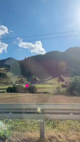 This video captures a scenic view of rolling green hills and mountains under a bright blue sky with scattered white clouds. The sun is shining, casting light and shadows across the landscape, with visible lens flares. In the foreground, there is a guardrail and dry grasses. The middle ground shows a field with scattered trees and some farm buildings, including a barn-like structure with a red roof. There are also what appear to be ski slopes or hiking trails on the mountainside. A single telephone pole stands prominently in the middle ground, and power lines run across the top of the frame. The camera appears to be moving, likely from inside a vehicle, as suggested by the perspective and the presence of the guardrail.
