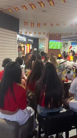 The video shows a crowded indoor space, possibly a bar or restaurant, with many people facing a television screen. The room is decorated with numerous small Spanish flags strung across the ceiling. The people are predominantly young adults, many wearing red and yellow clothing, indicating support for a sports team, likely the Spanish national team. Some individuals are holding noisemakers, suggesting an active and celebratory atmosphere. Several tables are visible, laden with drinks and bottles, and some people are seated while others are standing. The television screen displays a soccer match. The overall impression is of a group of fans watching a sporting event together.