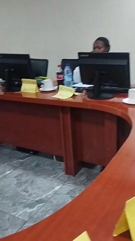Here is a detailed description of the video:

The video shows a conference room setting. A long, dark-wood table spans the length of the room, lined with computer monitors, keyboards, water bottles, and notebooks. People are milling around at the far end of the table. On the wall, a large screen displays a presentation titled "LAFARGE Safety Culture." One person is on the phone, wearing an orange safety vest and hard hat, while others stand nearby. The room has a grey tiled floor and light-colored walls. The overall atmosphere suggests a workplace environment where a presentation or meeting is taking place.