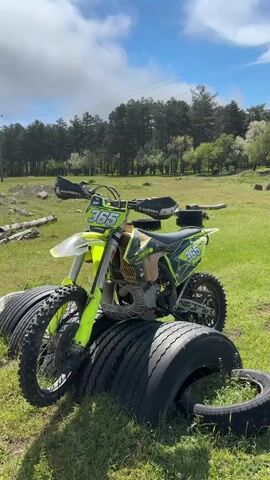 The video shows a lime green dirt bike parked in a grassy field. The bike has the number 365 on the front and sides. It is surrounded by stacks of tires. Trees are visible in the background under a partly cloudy sky. The camera focuses in on the number plate.