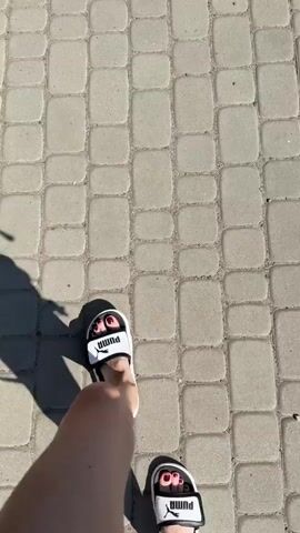 THE VIDEO SHOWS A FIRST-PERSON PERSPECTIVE OF SOMEONE WALKING ON A PAVED PATHWAY. THE PATHWAY IS MADE OF RECTANGULAR GRAY PAVING STONES. A SHADOW OF THE PERSON'S LEGS AND FEET IS VISIBLE ON THE RIGHT SIDE OF THE FRAME. THE PERSON IS WEARING SANDALS. THE SUN IS BRIGHT, CASTING A SHARP SHADOW.