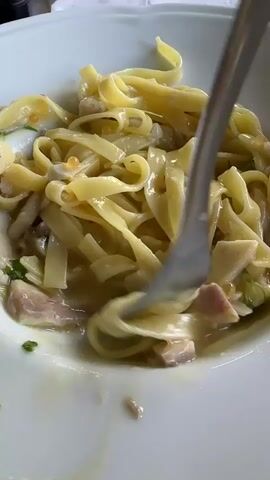 A fork is twirled into a bowl of fettuccine pasta, lifting a portion of the noodles and some pieces of fish and roe. The pasta is in a white bowl and appears to be in a creamy sauce.