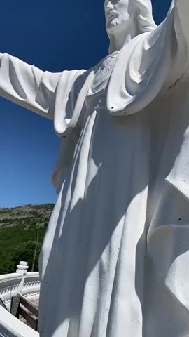 A close-up, low-angle shot of a large white statue of Jesus Christ, with his arms outstretched. The statue is made of concrete and is visible from the waist up. The sky is a clear, bright blue. In the background, there are green hills and a glimpse of a white railing. The sun is shining, casting shadows on the statue's robe. The camera appears to be handheld and pans slightly upwards, following the statue's form.