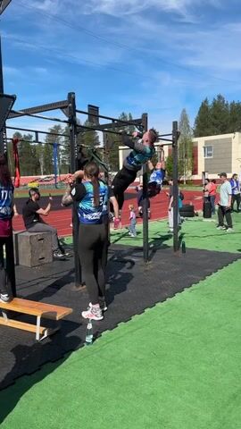The video shows a group of people participating in a fitness competition outdoors. They are performing pull-ups on a multi-station pull-up bar structure. The structure is black and labeled with "YOUSTEEL".  Some competitors are wearing weighted vests.  Several people are watching, some are filming with phones, and others are standing around the workout area. The setting includes a green artificial turf surface, a red running track, trees, and a building in the background under a blue sky with some clouds.