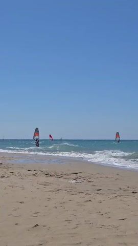 The video shows a beach scene with windsurfers in the ocean. The water is turquoise and there are small waves. The sand on the beach is light brown. In the foreground, a man is lying on his stomach on the sand, looking towards the ocean. In the background, there are several windsurfers with colorful sails on the water. Further in the distance, there is a rocky, arid coastline under a clear blue sky. The camera pans slowly across the scene.