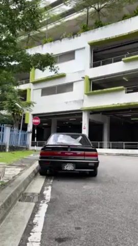The video shows a black Honda Accord parked near a parking garage. The car is facing away from the camera, and its license plate reads "JDD 3490". The parking garage is a multi-story structure with a white facade and green accents. There are trees and bushes visible in the background, and a street lamp and a stop sign are located near the car. The video is taken during the day.
