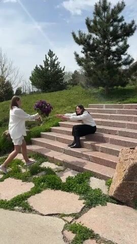 The video shows three different outdoor scenes. First, two women interact on a set of stone stairs. One woman in a white shirt and white shorts stands and hands a bouquet of purple flowers to a woman sitting on the stairs, wearing a white top and black bottoms. The second scene shows the woman in the white shirt and shorts walking away from the stairs, along a path made of large stone slabs. A third woman carrying a black bag bends down to pick something up from the ground near the stone path. The third scene shows the first woman walking on the stone path toward a lake. There are streetlights and trees along the path.