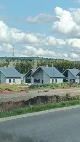 The video shows a row of modern, gray, unfinished houses with dark roofs. The houses are constructed with concrete walls and appear to be in a development or construction phase, with dirt mounds and some greenery visible around them. In the foreground, a paved road is visible, with a grassy verge and a metal guardrail. In the distance, a forest line is visible under a cloudy sky. A silver car is parked in front of one of the houses. A tall telecommunications tower is visible in the far distance. The camera appears to be mounted on a moving vehicle, providing a panning view of the houses and the surrounding landscape.