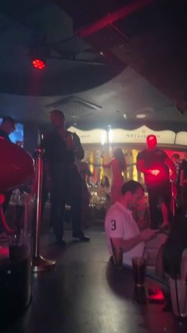 The video shows a busy bar with a lively atmosphere. Several people are visible, including bartenders, servers, and customers. Some are standing around the bar, others are seated at tables. A woman is holding a bottle with sparklers attached, with other women nearby. A DJ is playing music at a mixer with red lights. The environment is dimly lit with colorful lights, creating a club-like setting. Moet & Chandon branding can be seen in the background.