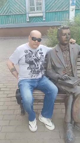 A man is sitting on a bench next to a bronze statue of a man holding a book. The man is wearing a grey t-shirt with a graphic print, blue jeans, and white sneakers. He has sunglasses on and his left arm is resting on the back of the bench. The statue is dressed in a suit and tie. The bench is made of wood and is situated on a paved area. Behind the bench is a wooden building with green trim and white window frames, and some greenery. The video captures the scene from a medium shot, with the camera slightly angled from the front.