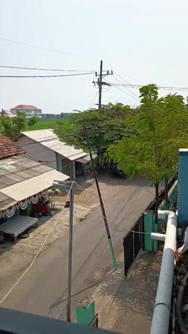 A high-angle medium shot shows a residential street lined with houses and trees under a clear, bright sky. The houses have pitched roofs, some with corrugated metal and others with red tiles. Several of the houses appear to be shops or market stalls, with awnings and displays visible. A utility pole with wires runs across the frame, leaning slightly to the right. The street is paved and empty except for a parked motorcycle. The overall impression is a quiet, sunlit day in a somewhat modest neighborhood.
