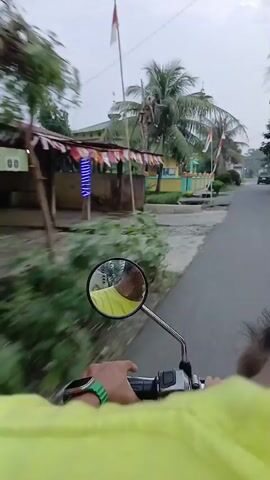 The video shows a first-person perspective of someone riding a motorcycle or scooter. In the foreground, the rider's hand with a green wristband and a watch is visible gripping the handlebar. The motorcycle's side mirror reflects the rider's face, wearing a yellow shirt. The environment appears to be a rural or suburban area with buildings, palm trees, and flags. Specifically, there's a building with yellow and turquoise walls, decorated with what looks like the Indonesian flag. Further down the road, another building with a dome and minaret, possibly a mosque, is visible. There's also a tall tower with lights. A red scooter with two people on it is seen passing by in the opposite direction. The road surface is somewhat rough, with some areas appearing to be dirt or gravel. The sky is overcast. The overall impression is of a casual ride through a tropical or subtropical region.